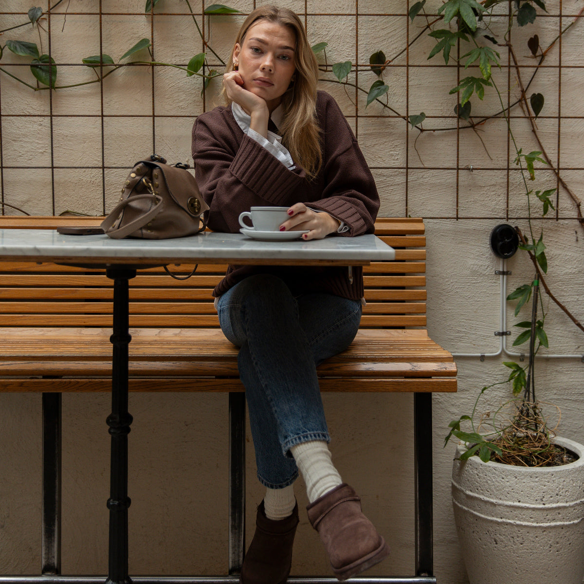 Woman sitting at a table with a cup of coffee in a cozy indoor setting with plants and a tiled wall.